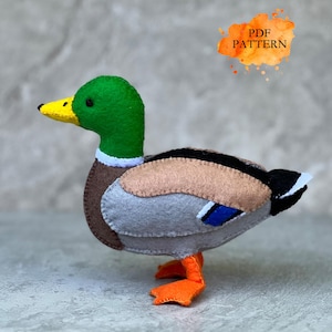 May include: A handmade felt duck with a green head, yellow beak, and orange feet. The duck's body features brown, tan, gray, and black felt. The image includes the text "PDF PATTERN" in an orange watercolor-style splash.