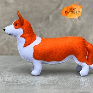 May include: A felt corgi dog toy in orange and white. The soft toy features a detailed design, with the text "PDF PATTERN" in an orange watercolor-style splash. The corgi is standing on a neutral surface.