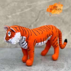 Puede incluir: Un tigre de peluche naranja con rayas negras y detalles blancos. El juguete tiene una cola con punta negra y se apoya en cuatro patas naranjas. La imagen incluye el texto "PDF PATTERN" en un salpicadura de estilo acuarela naranja.