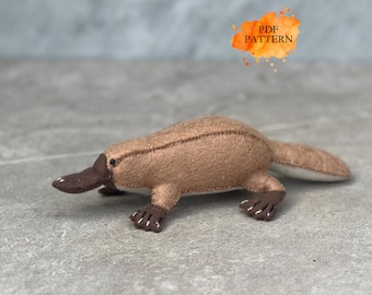 May include: A handmade felt platypus toy in shades of brown, with a dark brown bill and feet. The toy has a stitched design and the text "PDF PATTERN" in an orange watercolor splash.