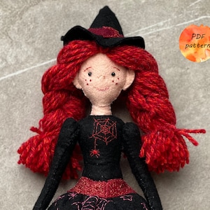 May include: A felt witch doll with red yarn hair, a black dress with a spiderweb design, and a black witch hat. The doll has a pink face with black eyes and freckles.
