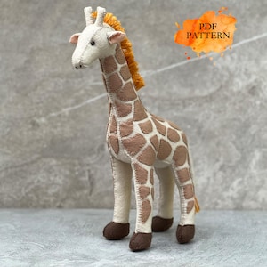 Könnte beinhalten: Ein handgefertigtes Giraffen-Spielzeug aus Filz mit cremefarbenem Kopf und Körper, braunen Flecken, dunkelbraunen Hufen und einer strukturierten orangefarbenen Mähne. Der Text PDF PATTERN ist schwarz auf einem orangefarbenen Aquarellfleck gedruckt.