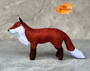 May include: A handmade felt fox figurine in shades of red, white, and brown. The fox has a white chest and tail tip, with dark brown legs and ears. The image includes the text "PDF PATTERN" in an orange watercolor-style splash.