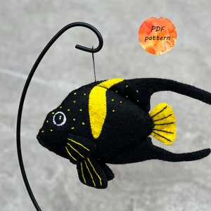 May include: A handmade felt fish ornament in black and yellow, suspended from a black metal stand. The fish features yellow accents on its head, fins, and tail, with small yellow dots. The ornament has the text "PDF pattern".
