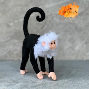 May include: A handmade monkey doll with a black body, white head, and a curled tail. The monkey has pink hands and feet. The text "PDF PATTERN" is in an orange splash in the upper right corner.
