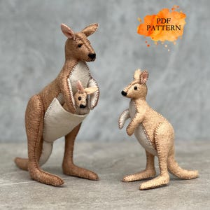 Puede incluir: Dos figuras de fieltro de canguro. Una es un canguro más grande con una cría en su bolsa. La otra es un canguro más pequeño de pie. Los canguros son de color marrón claro con vientres color crema y detalles cosidos. La imagen incluye el texto "PDF PATTERN".
