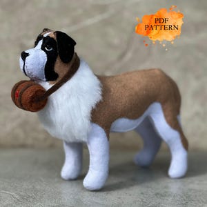 May include: A stuffed Saint Bernard dog with brown, white, and black markings, wearing a brown collar with a small barrel. The dog has white paws and a white chest. The image includes the text "PDF PATTERN".