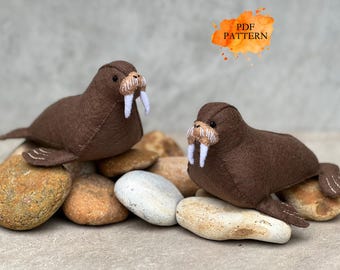 May include: Two brown felt walrus plush toys with white tusks and whiskers, resting on a bed of stones. The text "PDF PATTERN" is in an orange watercolor splash.