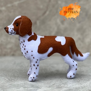 Peut inclure: Une figurine de chien en feutrine blanche avec des taches marron sur le corps, les pattes et le poitrail. La tête est marron avec une ligne blanche, un nez noir et des yeux sombres. Un motif aquarelle orange affiche le texte PDF PATTERN.