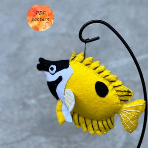 May include: A handmade felt fish ornament in yellow, black, and white, suspended from a black metal stand. The fish has a distinctive design with a black spot and a spiky dorsal fin. A watercolor circle with the text "PDF pattern" is in the background.