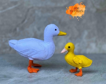 Puede incluir: Dos juguetes de pato de fieltro. Uno es un pato azul claro con patas naranjas y un pico amarillo. El otro es un pato amarillo más pequeño con patas naranjas y un pico rosa. El texto "PDF PATTERN" está en una forma de acuarela naranja.