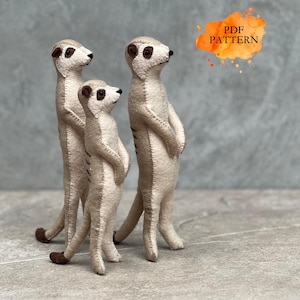 Op de afbeelding: Drie handgemaakte viltfiguren van stokstaartjes in staande houding, met lichtbeige lichamen, donkerbruine accenten rond de ogen en staartpunten, zichtbare stiksels langs de naden en een oranje grafische afbeelding met de tekst PDF PATTERN.