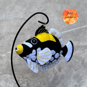 May include: A handmade felt fish ornament in black, yellow, and white, suspended from a black metal hook. The fish features detailed stitching and a whimsical design. A small orange circle with the text "PDF pattern" is in the background.