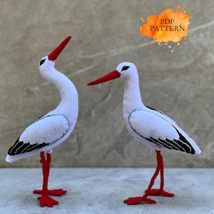 Puede incluir: Dos figuras de fieltro blanco de cigüeñas con picos y patas rojas. Las cigüeñas tienen detalles negros en sus alas y están de pie sobre una superficie clara. La imagen incluye el texto "PDF PATTERN".