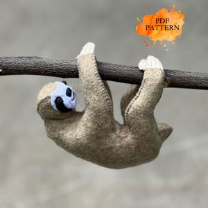 Peut inclure: Un jouet paresseux en feutre suspendu à une branche brune. Le paresseux est brun clair avec un visage blanc et noir et des griffes blanches. L'image comprend le texte "PDF PATTERN" dans une éclaboussure d'aquarelle orange.
