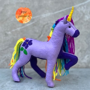 Peut inclure: Licorne en feutre violet avec une crinière et une queue arc-en-ciel. La licorne a une fleur brodée sur le côté et une corne dorée. Le texte "PDF pattern" est écrit sur un fond aquarelle.