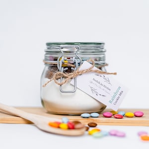 May include: A glass jar filled with cookie mix and colourful sprinkles, tied with twine and a tag that reads "Norfolk Jar Kitchen After Hours Bake Sale" and "Rainbow Cookie Mix".