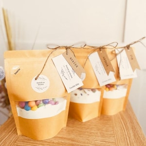 May include: A collection of kraft paper bags containing cookie mix and sweets. Each bag has a clear window showing the contents. The bags are tied with twine and labelled with tags reading "Winter", "Arthur", and "Alfie". The bags are on a wooden surface.
