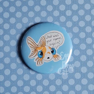 Op de afbeelding: Een blauwe knop met een cartoon-goudvis met oranje en witte schubben en blauwe ogen. De goudvis zegt "Hoe heette je nog een keer?" in een spreekballon.