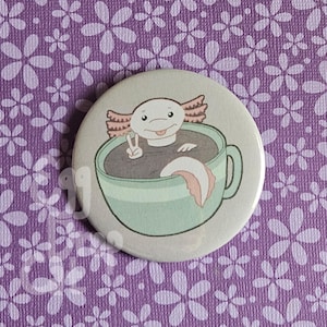 Op de afbeelding: Een witte axolotl met roze kieuwen zit in een lichtblauwe theekop met een bruine vloeistof. De axolotl geeft het vredesteken met zijn voorpoten. De theekop heeft een wit handvat.