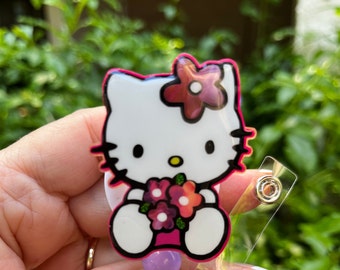 May include: A pink Hello Kitty character charm with a white face, pink bow, and a bouquet of flowers. The charm is attached to a clear plastic badge holder with a metal ring.