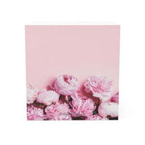 Puede incluir: Un fondo rosa con una colección de peonías rosas en la parte inferior. Las flores, con sus pétalos suaves y en capas, están en varias etapas de floración. El texto "Pretty Pink Paperie Co." es visible.