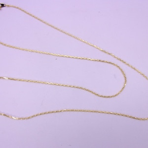 May include: A delicate gold-colored necklace with a fine chain. The necklace is laid out on a light-colored surface, showcasing its length and design. The chain has a subtle, textured appearance, and a clasp is visible at one end.