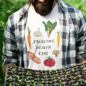 May include: A white t-shirt with the text "PROACTIVE HEALTH CARE" and vegetable illustrations. The person is wearing a plaid shirt and holding a tray of seedlings in a greenhouse.