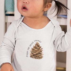 May include: A baby wearing a white long-sleeved bodysuit with the text "Enjoy the little things" and a brown pine cone graphic. The baby is standing on a striped rug.