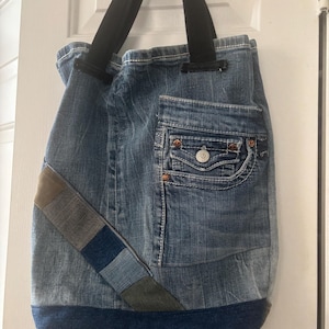 Può includere: Borsa tote in denim riciclato con cinghie nere. La borsa presenta un design patchwork con varie tonalità di denim blu e una base blu scuro. Una tasca in denim è visibile.