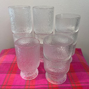 May include: A set of ten clear glass drinking glasses with a textured surface. The glasses are stacked in two piles of five. The glasses are on a pink and red plaid tablecloth.