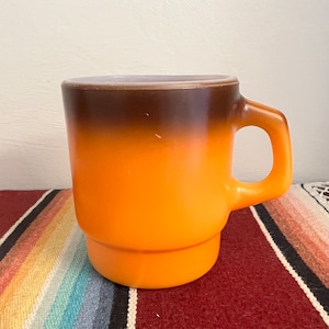 May include: A brown and orange ombre mug with a white rim. The mug has a rounded handle and a stacked base.