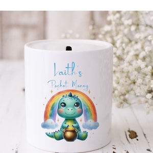 May include: White ceramic money bank with a cartoon dinosaur design. The bank features a rainbow, clouds, and the text "Laith's Pocket Money" in blue script. The dinosaur is holding a pot of gold.