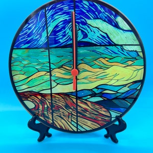 May include: A stained glass clock with a blue, green, and yellow design. The clock is on a black stand and has a red hour hand.