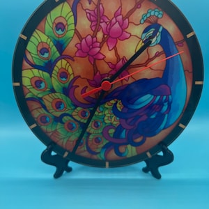 May include: A round clock with a peacock design. The clock face features a stained glass style peacock with a blue body, green and yellow feathers, and pink flowers. The clock hands are black with a red second hand.