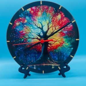 May include: A round clock featuring a colourful tree design. The clock face is black with gold accents and the tree is a vibrant mix of red, blue, yellow, and green. The clock is on a black stand.