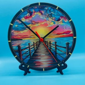 May include: A round clock with a colourful stained glass design of a sunset over a wooden pier extending into the ocean. The clock has black hands and a gold rim.