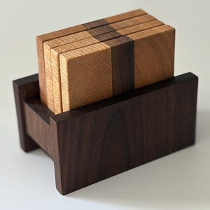 May include: A set of four rectangular wooden coasters featuring a two-tone design with light oak and dark walnut wood grain, held upright in a matching dark brown wooden storage stand with a rectangular base and two side supports.