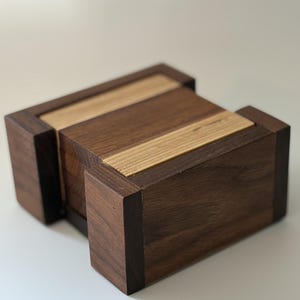 May include: A rectangular wooden box constructed from alternating dark walnut and light oak wood panels. The design features a central section with horizontal wood grain patterns flanked by two solid dark wood end blocks, creating a geometric structure.