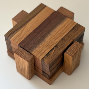 May include: A stack of square wooden coasters featuring alternating light and dark brown wood grain patterns. The coasters are held together by a matching wooden frame with four protruding rectangular supports on each side of the square base.