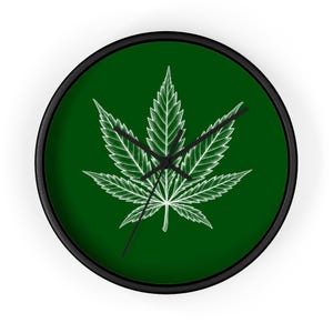 May include: A round wall clock with a black frame and a dark green face. The center of the clock features a white, detailed illustration of a seven-pointed cannabis leaf. Three thin black hands are positioned in the center of the clock face.