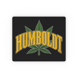 May include: A rectangular black mouse pad featuring a green cannabis leaf graphic centered behind the word HUMBOLDT written in large, bold, yellow capital letters with a slight arch and a thin white outline against the dark background.