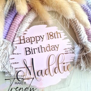 May include: A clear, hexagonal cake topper with the text "Happy 18th Birthday Maddie" in black and gold lettering. The topper is set against a pink background with a brushstroke design, and a macrame rainbow.
