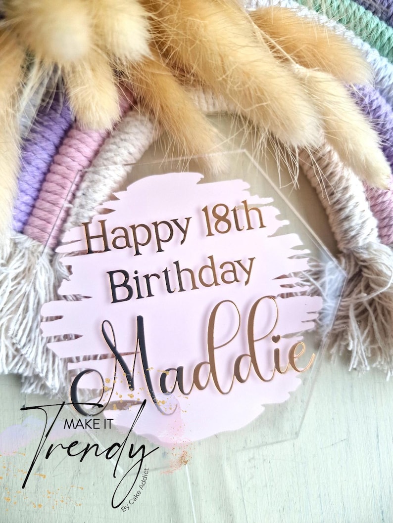 May include: A clear, hexagonal cake topper with the text "Happy 18th Birthday Maddie" in black and gold lettering. The topper is set against a pink background with a brushstroke design, and a macrame rainbow.