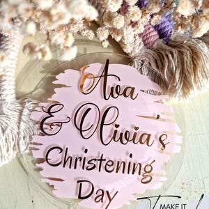 May include: A clear acrylic cake topper with the text "Ava & Olivia's Christening Day" in black script over a pink brushstroke design. The word "Ava" is in gold script. The background includes a macrame wall hanging and dried flowers.