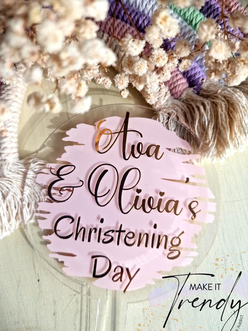 May include: A clear acrylic cake topper with the text "Ava & Olivia's Christening Day" in black script over a pink brushstroke design. The word "Ava" is in gold script. The background includes a macrame wall hanging and dried flowers.
