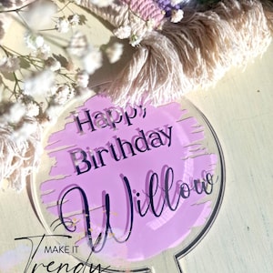 May include: A clear acrylic cake topper with the words "Happy Birthday Willow" in black script over a light purple background. The words "Make it Trendy" are also visible. The background includes a rainbow macrame.