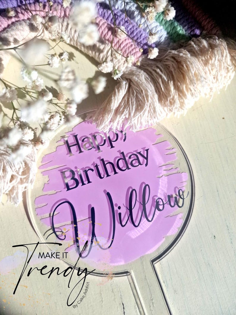 May include: A clear acrylic cake topper with the words "Happy Birthday Willow" in black script over a light purple background. The words "Make it Trendy" are also visible. The background includes a rainbow macrame.