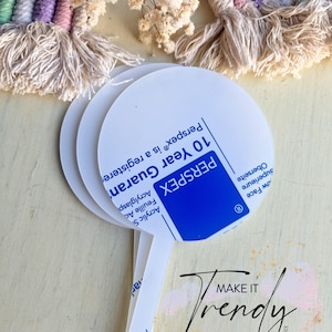 May include: White Perspex cake toppers with a round top and a handle. The toppers have the text "10 Year Guarantee" and "Perspex" printed on them. The image also includes a rainbow macrame and dried flowers.