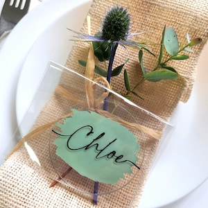May include: A clear acrylic place card with the name "Chloe" in black script, set on a burlap napkin. The card features a light green painted accent and is adorned with a thistle and greenery, placed on a white plate.